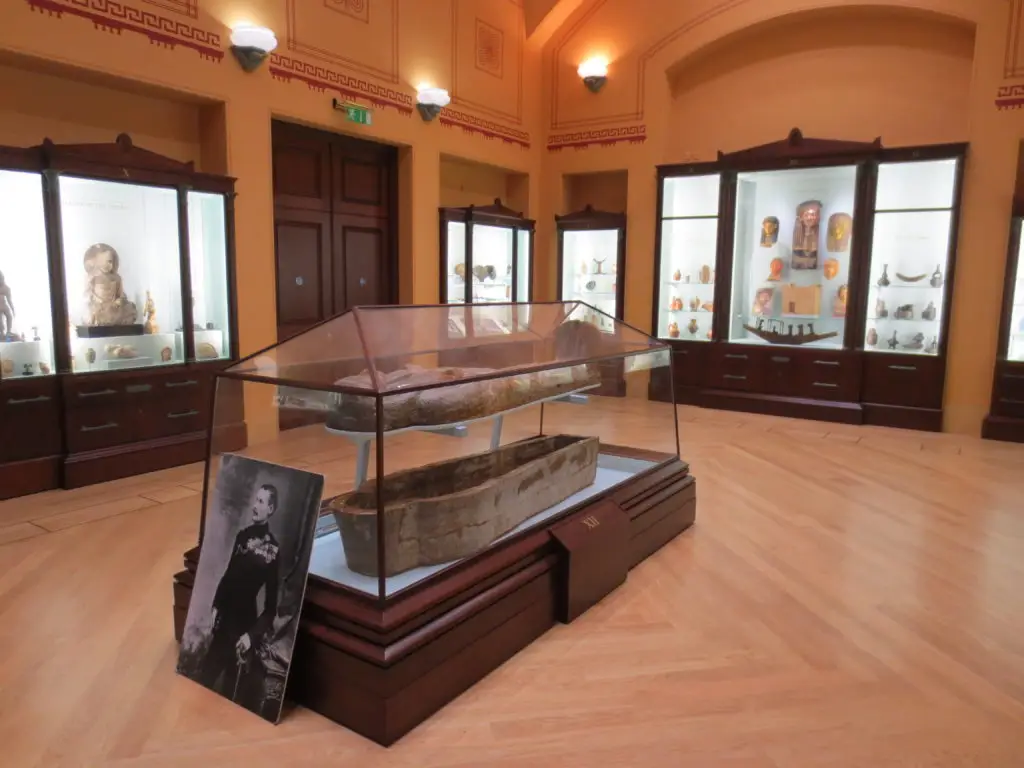 An Egyptian wooden coffin in a glass case in a neoclassical hall, a portrait photograph of an officer beside it